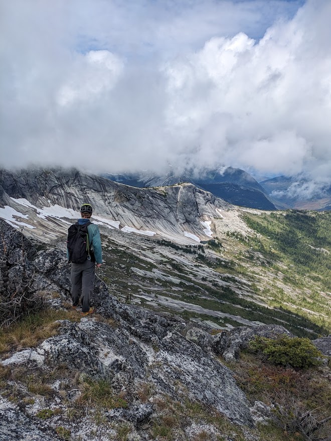 A climber on a rocky ridge looking across at long granite slabs and cliff bands with cloud sitting on the summits