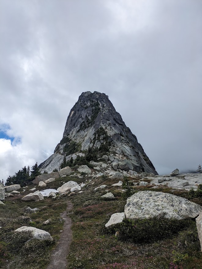The steep granite tower of Vicuna Peak rising above boulder-strewn subalpine meadows, with a faint trail leading toward it