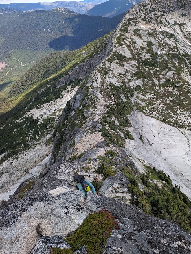 Looking down the ridge at a climber moving up the crack at the crux, with the valley far below